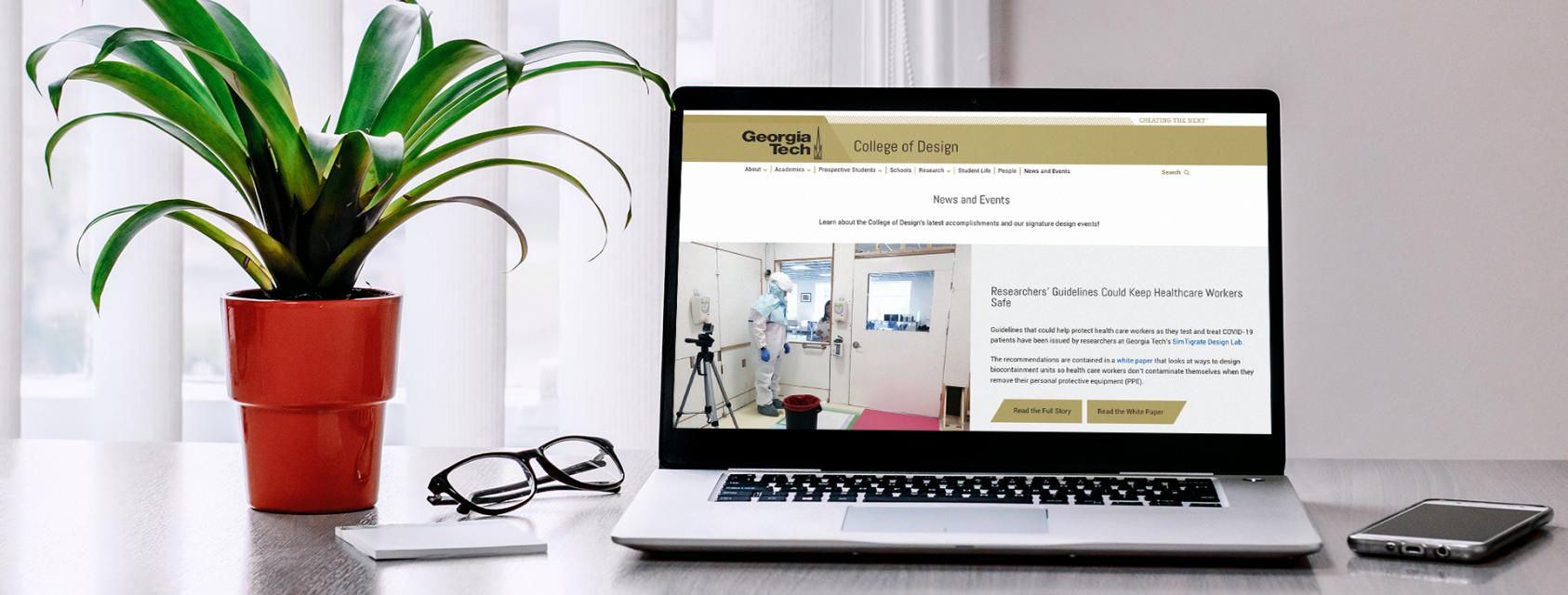 A computer showing the College of Design website sitting on a desk with glasses and a plant.