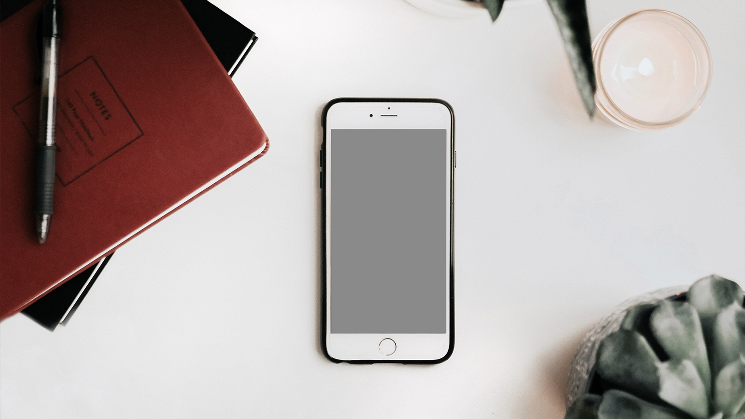 An iPhone sitting on a desk with a notebook and a plant, seen from the top.